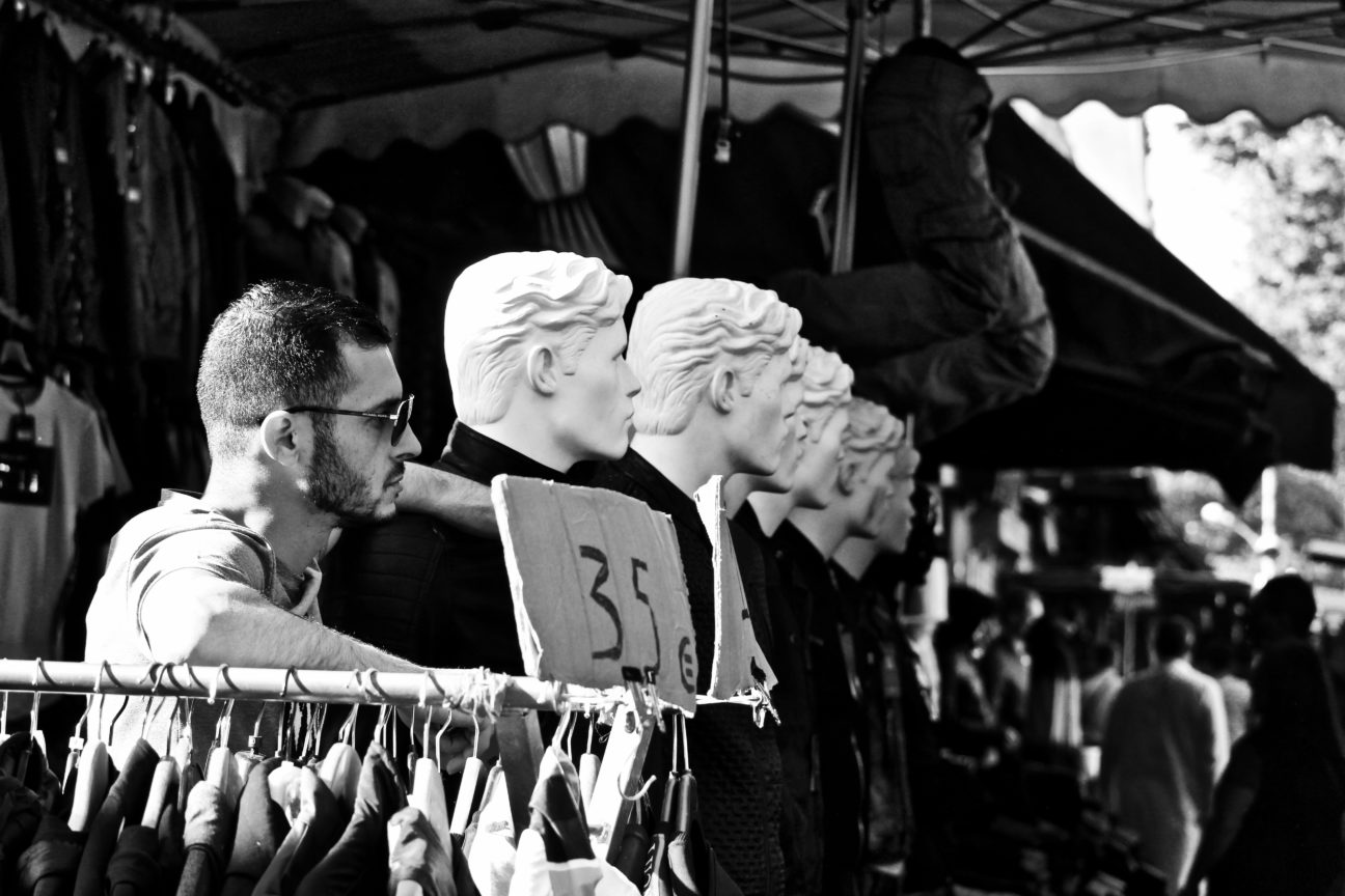 Photo en noir et blanc, prise en avril 2018 aux Puces de Saint-Ouen, du visage de profil d'un vendeur de vêtement, dont la tête finit un alignement des têtes de mannequins de celluloid qui semblent regarder tous au même endroit